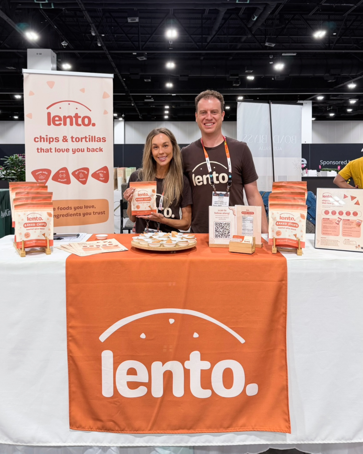 alex and travis smiling behind the lento. booth at newtopia now, holding a bag of baked chips